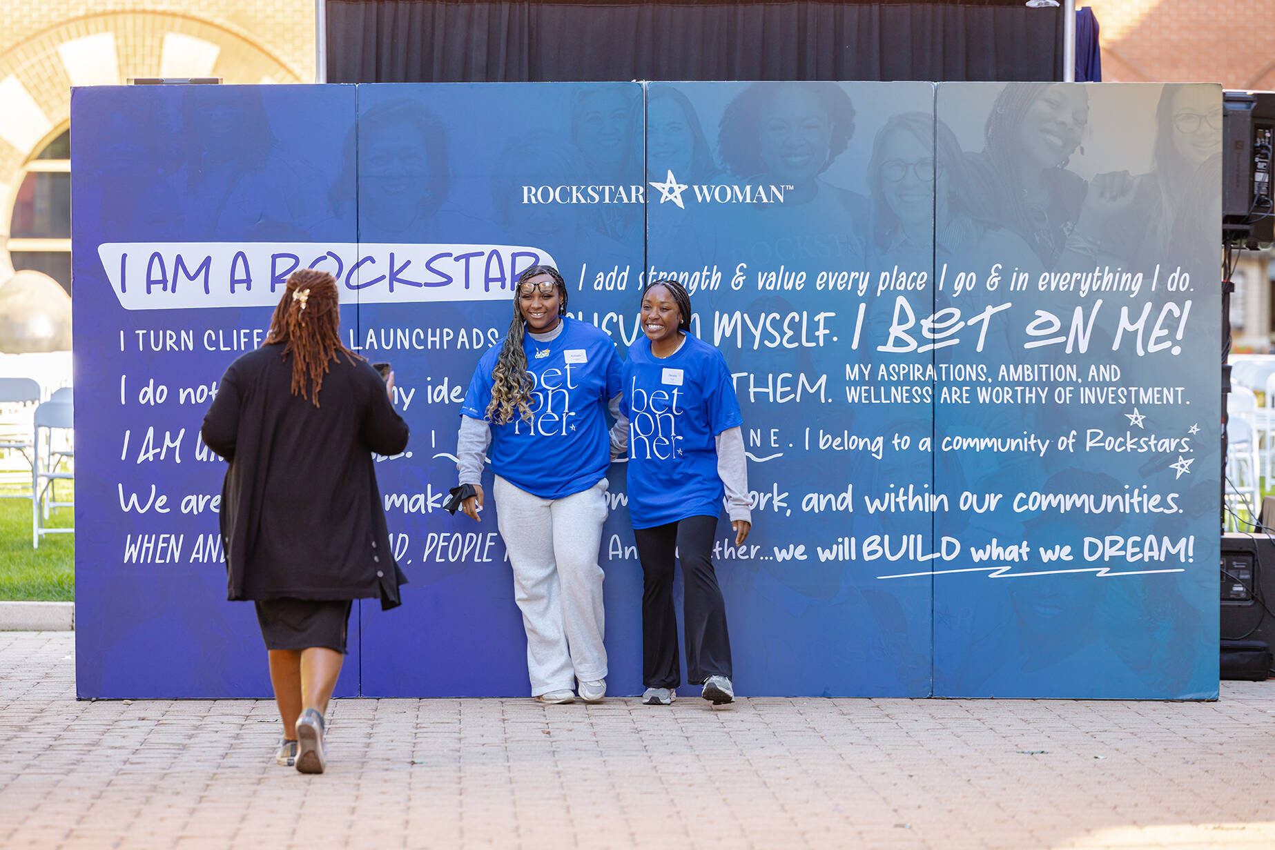 Two students in blue t-shirts stand against a backdrop created for a rockstar woman event on the Pew Grand Rapids campus while a person takes their photo